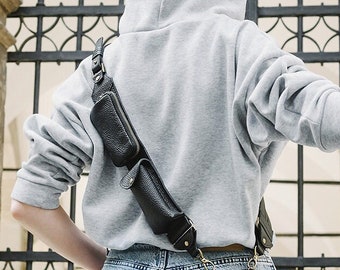 Può includere: Una custodia a tracolla per telefono in pelle nera dotata di una lunga tracolla regolabile con due piccole tasche integrate. La custodia è fissata alla tracolla con elementi in metallo e sostiene uno smartphone verticalmente sul fianco.