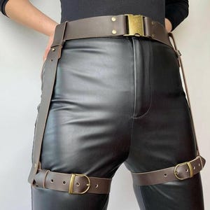 Può includere: Una cintura in pelle marrone con fibbia color oro e cinturini per le gambe abbinati. La cintura e i cinturini sono fissati con fibbie color oro. La persona indossa pantaloni di pelle neri e una maglia nera a maniche lunghe.
