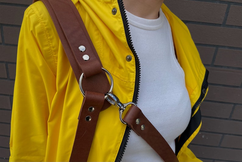 May include: A brown leather camera strap with silver hardware and two adjustable rings. The strap is worn over a yellow jacket.