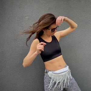 May include: A black sleeveless crop top paired with white and black patterned leggings and a white fanny pack featuring long fringe detailing. The fanny pack is worn around the waist with a zipper closure and a silver-toned metal buckle on the strap.