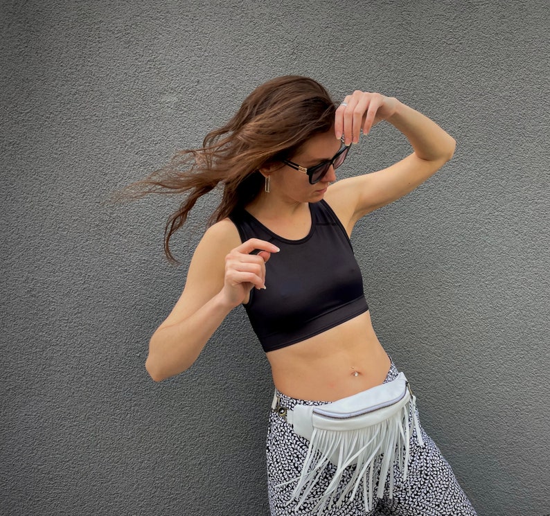 May include: A black sleeveless crop top paired with white and black patterned leggings and a white fanny pack featuring long fringe detailing. The fanny pack is worn around the waist with a zipper closure and a silver-toned metal buckle on the strap.