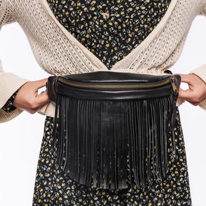 May include: Black leather fanny pack with a fringe detail and a gold zipper. The bag is worn around the waist. The person is wearing a cream-colored cardigan over a floral print dress.
