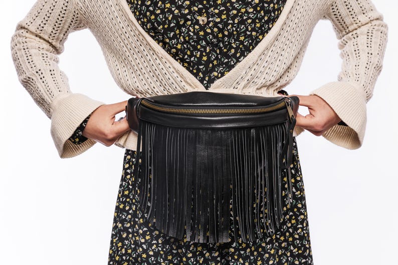 May include: Black leather fanny pack with a fringe detail and a gold zipper. The bag is worn around the waist. The person is wearing a cream-colored cardigan over a floral print dress.