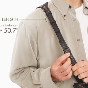 May include: A black textured leather strap with brass hardware and a buckle adjustment mechanism. The text on the image indicates a strap length adjustable between 46 inches and 50.7 inches. The strap is worn over a beige button-down shirt.