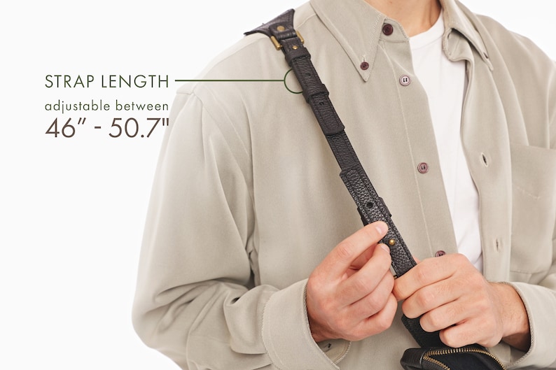 May include: A black textured leather strap with brass hardware and a buckle adjustment mechanism. The text on the image indicates a strap length adjustable between 46 inches and 50.7 inches. The strap is worn over a beige button-down shirt.