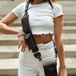 May include: A dark brown leather crossbody bag featuring a wide adjustable strap with two attached zippered pouches and a larger rectangular pocket at the base. The bag is worn diagonally across the torso, showcasing a textured finish and silver-toned hardware.