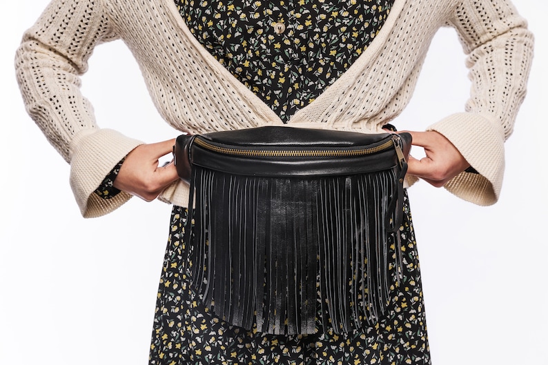 May include: A black leather fanny pack with a fringe detail. The fanny pack is worn around the waist of a person wearing a white cardigan and a floral dress.