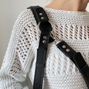 May include: Black leather camera strap with silver hardware and adjustable length. The strap is worn over a white crocheted sweater.