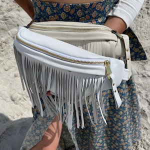 May include: Two fanny packs with long fringe detailing. The front bag is bright white with a gold-tone zipper, while the bag behind it is a cream-colored off-white shade. Both bags feature a smooth finish and adjustable straps with gold hardware.