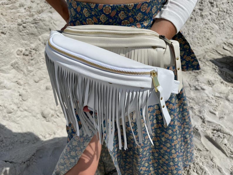 May include: Two fanny packs with long fringe detailing. The front bag is bright white with a gold-tone zipper, while the bag behind it is a cream-colored off-white shade. Both bags feature a smooth finish and adjustable straps with gold hardware.