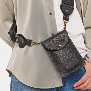 May include: A black textured crossbody phone pouch with a flap closure, a central snap button, and antique brass hardware. The pouch is attached to a wide, adjustable black strap that features matching textured detailing and metal rivets.