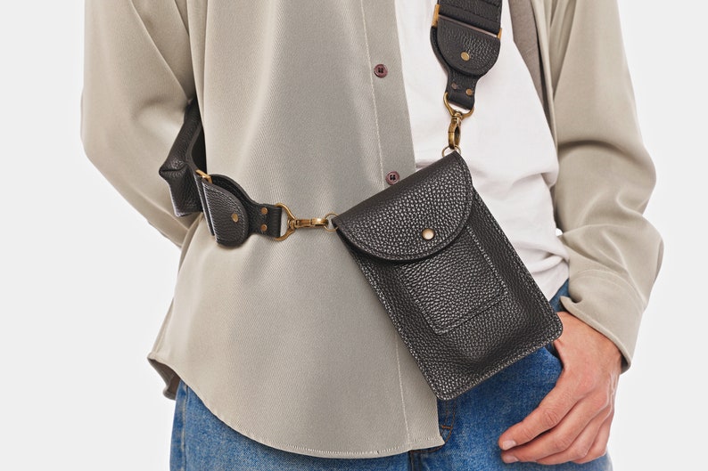 May include: A black textured crossbody phone pouch with a flap closure, a central snap button, and antique brass hardware. The pouch is attached to a wide, adjustable black strap that features matching textured detailing and metal rivets.