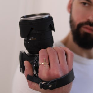 Può includere: Una fotocamera DSLR nera con un obiettivo e un cinturino da polso in pelle nera. Un anello d'oro con un design a croce è sulla mano che tiene la fotocamera. L'obiettivo ha la scritta "24-70mm 1:2.8 DG HSM".