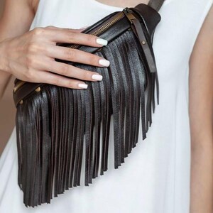 May include: Black leather bum bag with long fringe detail. The bag has a gold zip and gold studs along the top. The strap is adjustable. The bag is worn across the body.