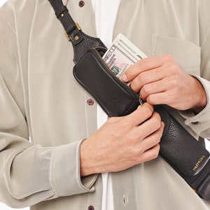 May include: A black textured leather crossbody sling bag with a zippered compartment containing a stack of US dollar bills. The bag features a long adjustable strap with metallic hardware and is worn diagonally across the chest over a beige shirt.