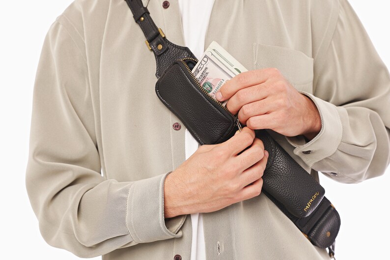 May include: A black textured leather crossbody sling bag with a zippered compartment containing a stack of US dollar bills. The bag features a long adjustable strap with metallic hardware and is worn diagonally across the chest over a beige shirt.