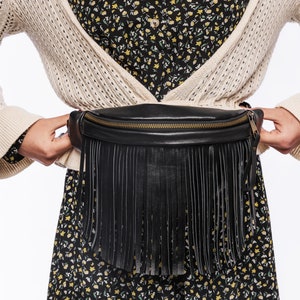 May include: A black leather fanny pack featuring a long fringe detail along the front panel and a gold-toned metal zipper closure. The bag is worn around the waist, showcasing a smooth texture and a structured, curved top edge design.