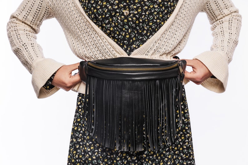 May include: A black leather fanny pack featuring a long fringe detail along the front panel and a gold-toned metal zipper closure. The bag is worn around the waist, showcasing a smooth texture and a structured, curved top edge design.