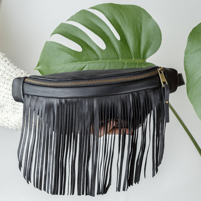 May include: Black leather fanny pack with long fringe detail and a gold zip. The bag is shown against a white background with a large green leaf.