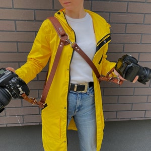 May include: A person wearing a yellow raincoat with a brown leather camera strap and two black cameras. The strap is attached to the cameras and has a metal ring on the front.