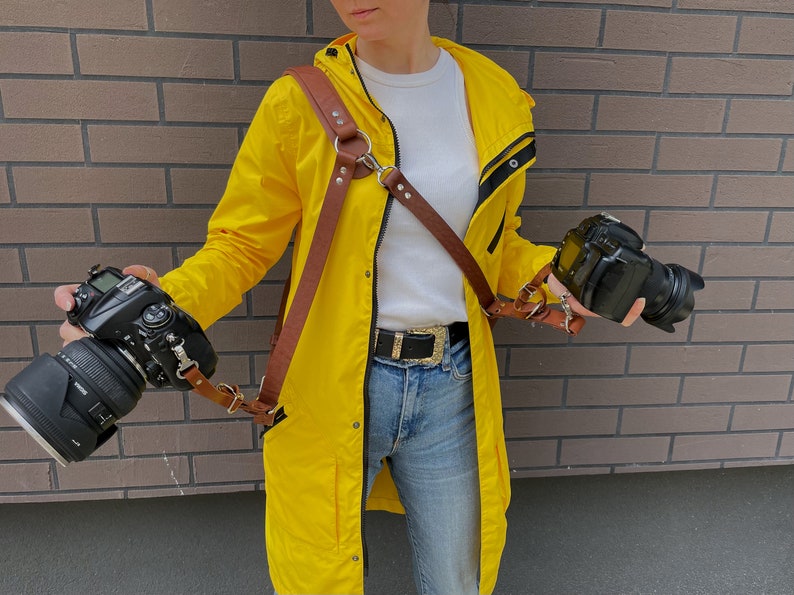 May include: A person wearing a yellow raincoat with a brown leather camera strap and two black cameras. The strap is attached to the cameras and has a metal ring on the front.
