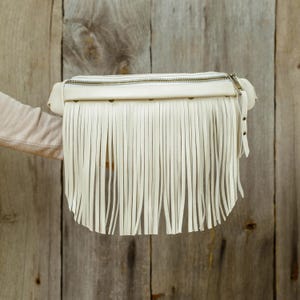 May include: Cream-coloured fanny pack with long fringe detail. The bag has a zip closure and a small strap with metal rivets. The bag is held against a wooden background.