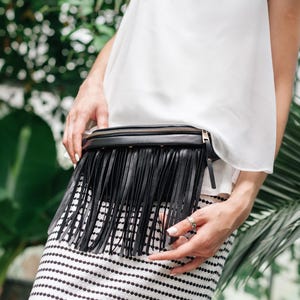 May include: Black leather belt bag with long fringe detail. The bag has a zip closure and is worn around the waist. The person in the image is wearing a white top and a black and white striped skirt. Green foliage forms the background.
