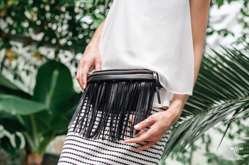 May include: Black leather belt bag with long fringe detail. The bag has a zip closure and is worn around the waist. The person in the image is wearing a white top and a black and white striped skirt. Green foliage forms the background.