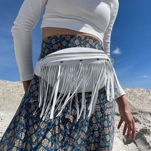 May include: A white fanny pack featuring long white fringe detailing along the front panel and a gold-toned zipper closure. The bag is worn around the waist over a blue skirt with a multi-colored floral pattern and a white long-sleeved ribbed top.