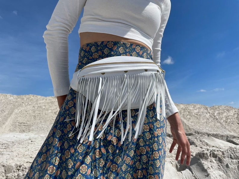 May include: A white fanny pack featuring long white fringe detailing along the front panel and a gold-toned zipper closure. The bag is worn around the waist over a blue skirt with a multi-colored floral pattern and a white long-sleeved ribbed top.