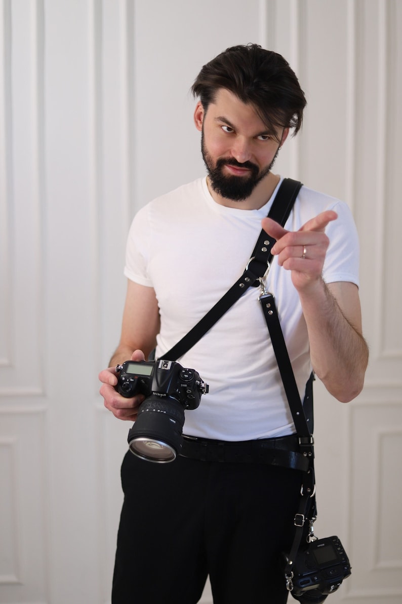 May include: A man wearing a white t-shirt and black trousers is holding a black camera with a lens attached. He is wearing a black leather camera strap with metal accents. The camera strap is worn across his body and has a second camera attached to it.