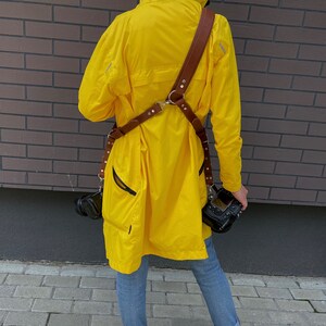 May include: A person wearing a yellow raincoat with a brown leather camera strap and two cameras attached. The person is wearing blue jeans and white sneakers.