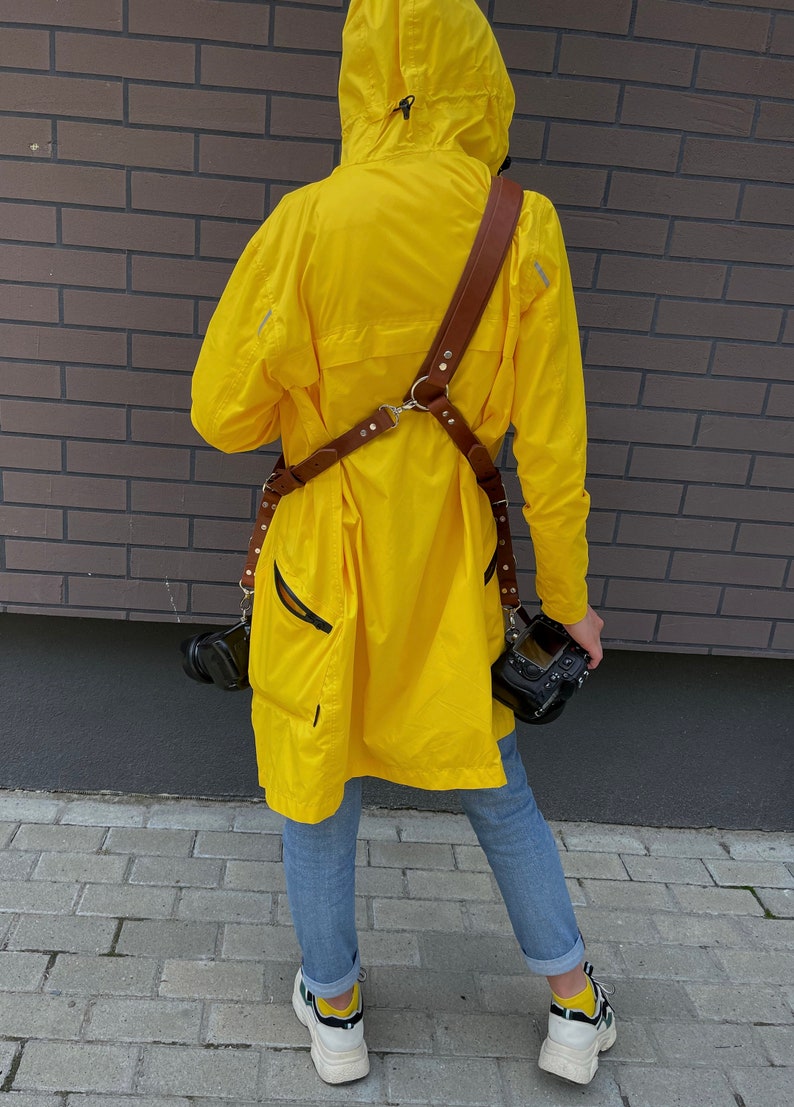 May include: A person wearing a yellow raincoat with a brown leather camera strap and two cameras attached. The person is wearing blue jeans and white sneakers.