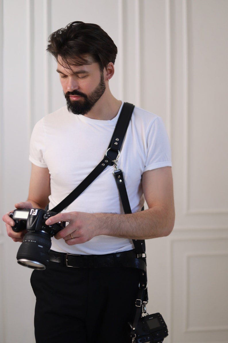 May include: A man wearing a white t-shirt and black pants is holding a black DSLR camera with a lens attached. He is wearing a black leather camera strap with a silver buckle.