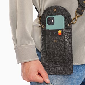 May include: A black pebbled leather crossbody phone pouch with a rounded flap, a snap closure, and an external card slot containing a credit card. The pouch features antique brass hardware, including D-rings and a matching adjustable shoulder strap.