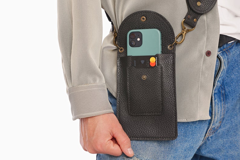 May include: A black pebbled leather crossbody phone pouch with a rounded flap, a snap closure, and an external card slot containing a credit card. The pouch features antique brass hardware, including D-rings and a matching adjustable shoulder strap.