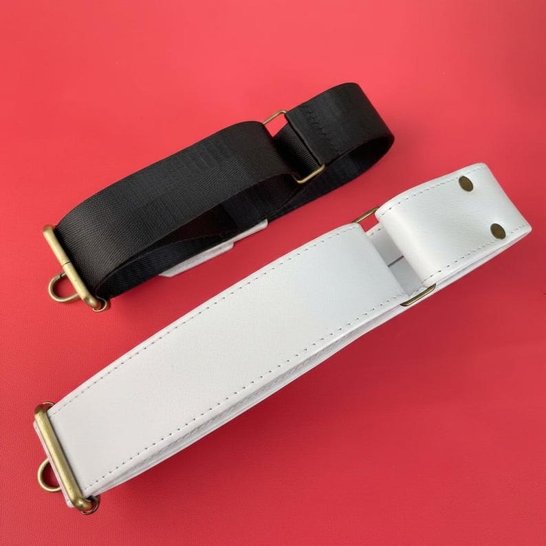 May include: Two bag straps, one black and one white, are displayed against a red background. The black strap is made of a woven material, while the white strap is leather. Both straps have gold-colored metal hardware, including D-rings and adjustable buckles.