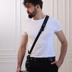 May include: A man wearing a white t-shirt and black pants with a black leather camera strap and a black camera.