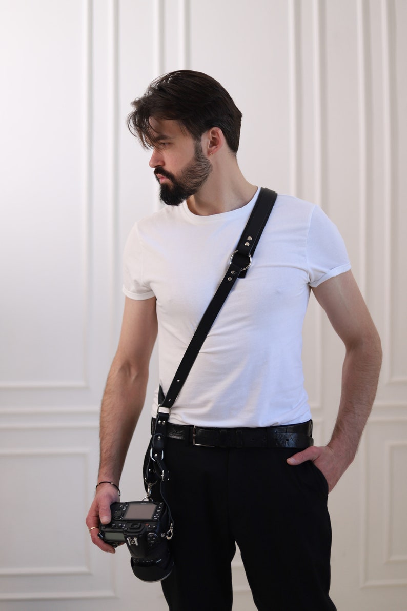 May include: A man wearing a white t-shirt and black pants with a black leather camera strap and a black camera.