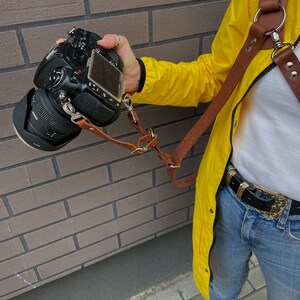 May include: A brown leather camera strap with a metal ring and buckle. The strap is attached to a black DSLR camera and is being worn by a person wearing a yellow jacket and blue jeans.