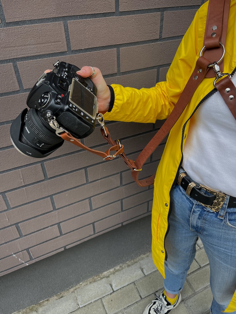May include: A brown leather camera strap with a metal ring and buckle. The strap is attached to a black DSLR camera and is being worn by a person wearing a yellow jacket and blue jeans.