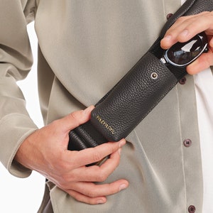May include: A dark brown, pebbled leather glasses case with a gold-tone metal snap closure and gold embossed lettering. The cylindrical case is designed to be worn as a crossbody accessory, featuring a matching leather strap and a secure flap top.