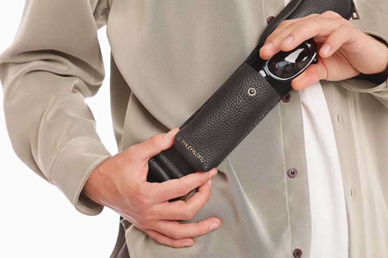 May include: A dark brown, pebbled leather glasses case with a gold-tone metal snap closure and gold embossed lettering. The cylindrical case is designed to be worn as a crossbody accessory, featuring a matching leather strap and a secure flap top.