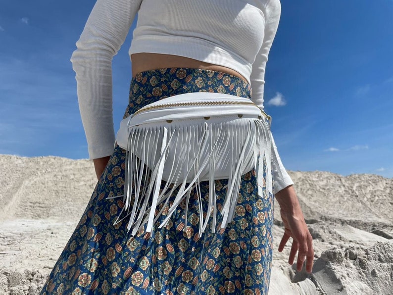 May include: White leather fanny pack with fringe detail and a gold zipper. The bag is worn around the waist and is paired with a floral print skirt and a white long-sleeve shirt. The background is a bright blue sky and sand.