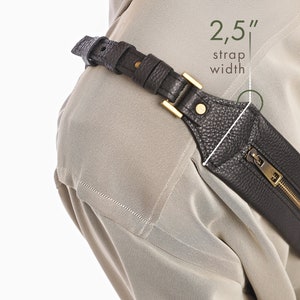 May include: A dark brown textured leather strap with a gold-tone metal buckle and a zippered pocket. The strap features a 2.5 inch strap width as indicated by the text overlay. The accessory is worn over a light beige garment with visible stitching.
