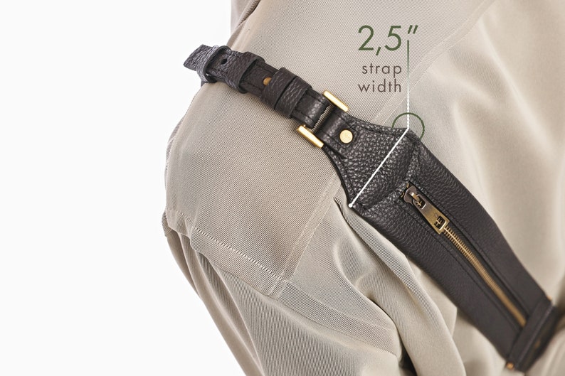 May include: A dark brown textured leather strap with a gold-tone metal buckle and a zippered pocket. The strap features a 2.5 inch strap width as indicated by the text overlay. The accessory is worn over a light beige garment with visible stitching.