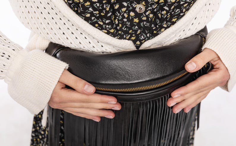 May include: Black leather fanny pack with a gold zip and fringe detail. The bag is worn around the waist. The person is wearing a cream-coloured sweater and a floral print top. The bag is held by the hands.