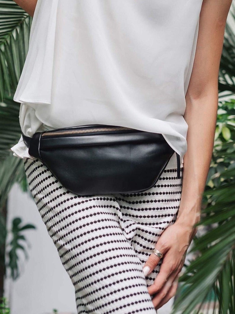 May include: A black leather fanny pack with a gold zipper. The fanny pack is worn around the waist of a person wearing a white top and black and white striped pants.