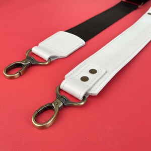May include: Two bag straps with metal clasps. One strap is white leather with brass-colored hardware and the other is black fabric. The straps are set against a red background.