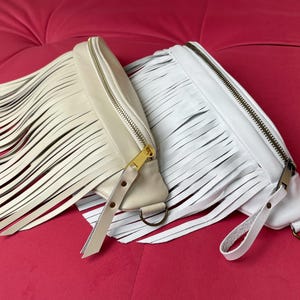 May include: Two fringed leather fanny packs in cream and white, with zip closures and strap details. The cream-coloured bag has a gold zip, while the white bag has a silver zip. Both bags have a D-ring attachment.